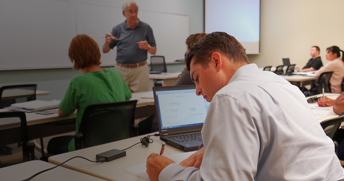 student taking notes while professor lectures at the front of class