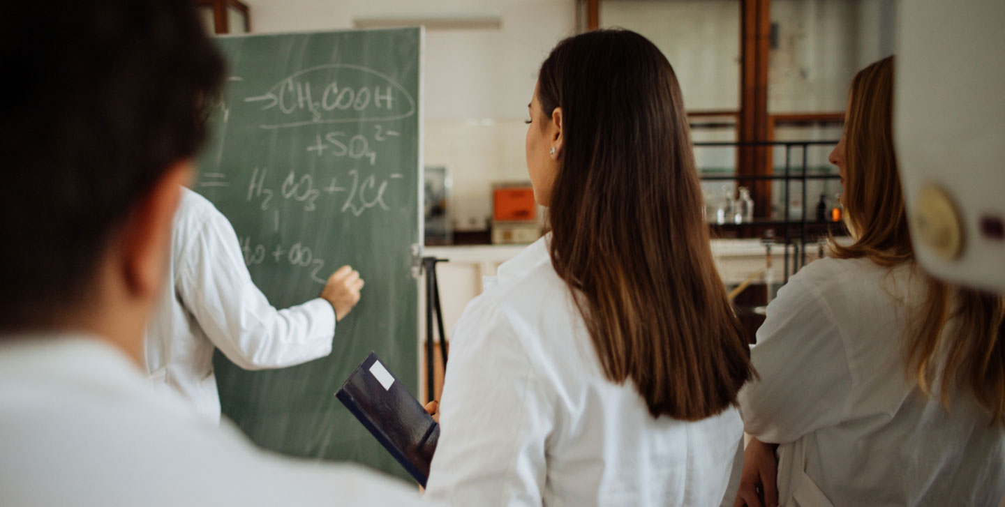 student facing a chalk board watching someone write chemical equations; chemistry