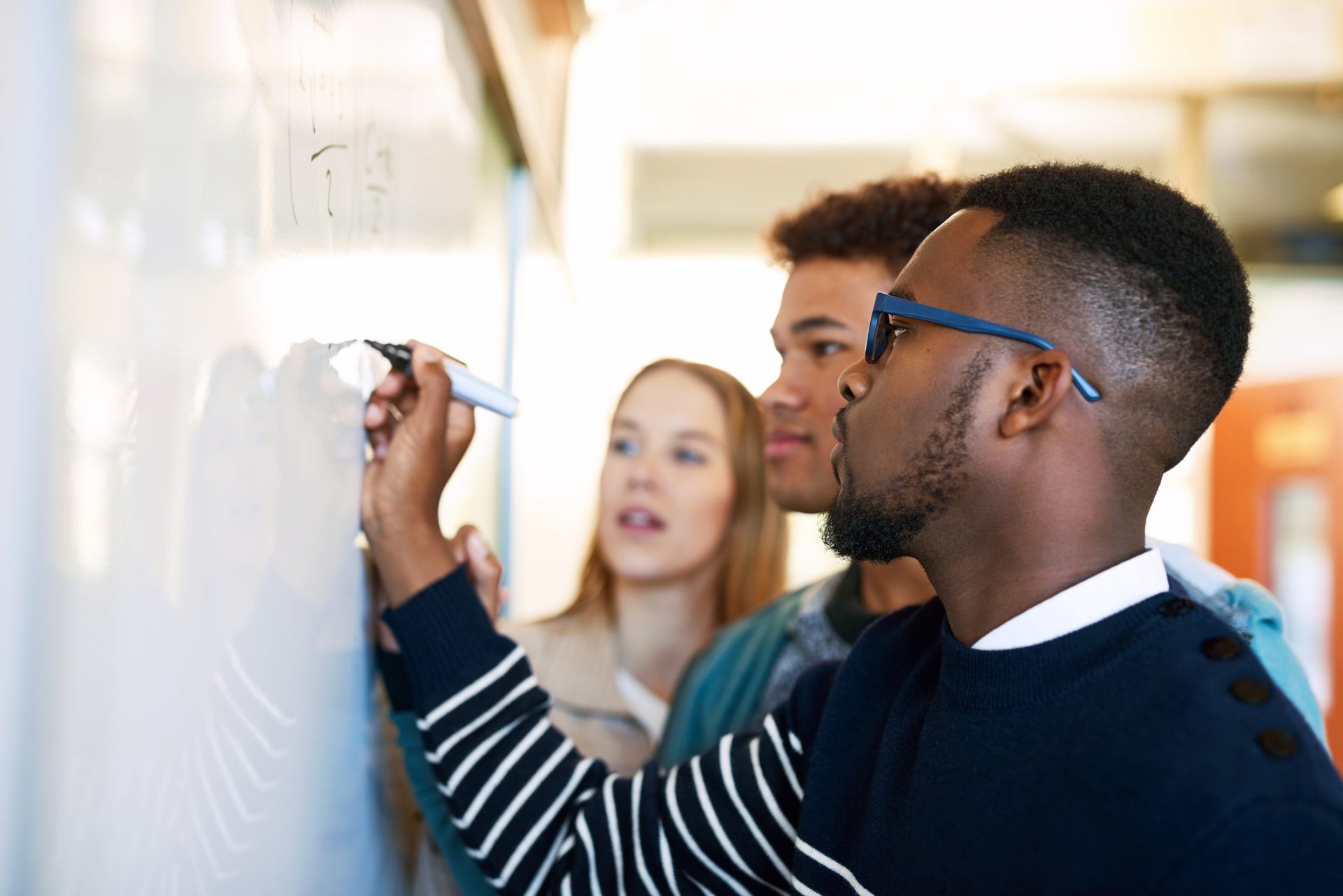 Shot of a young man writing on a whiteboard while students look on; mathematics
