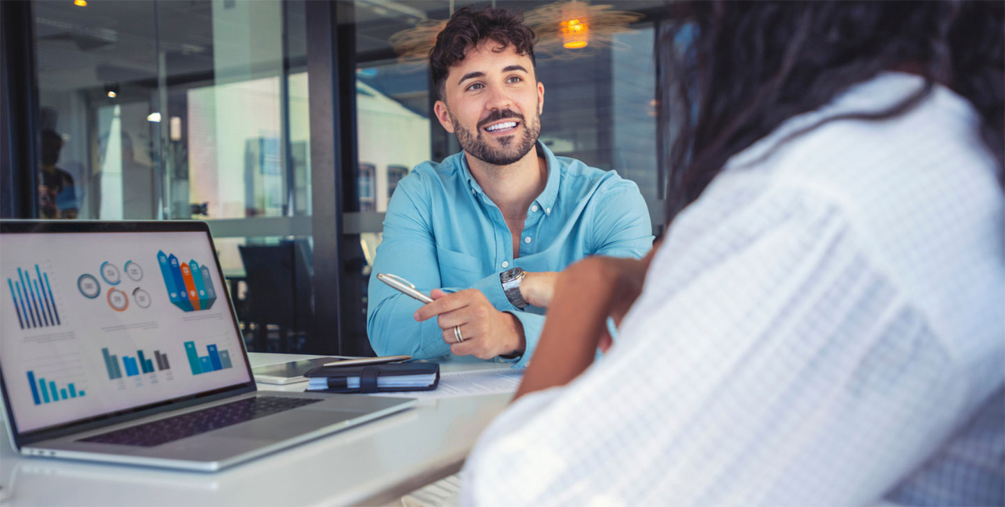 male student working with female at table with laptop with business graphs on screen