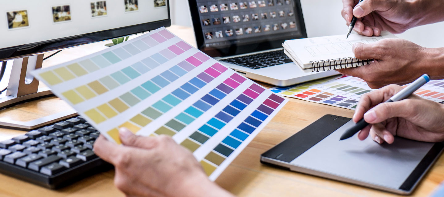 hand holding color chart while two people take notes, computer in background; graphic arts and design