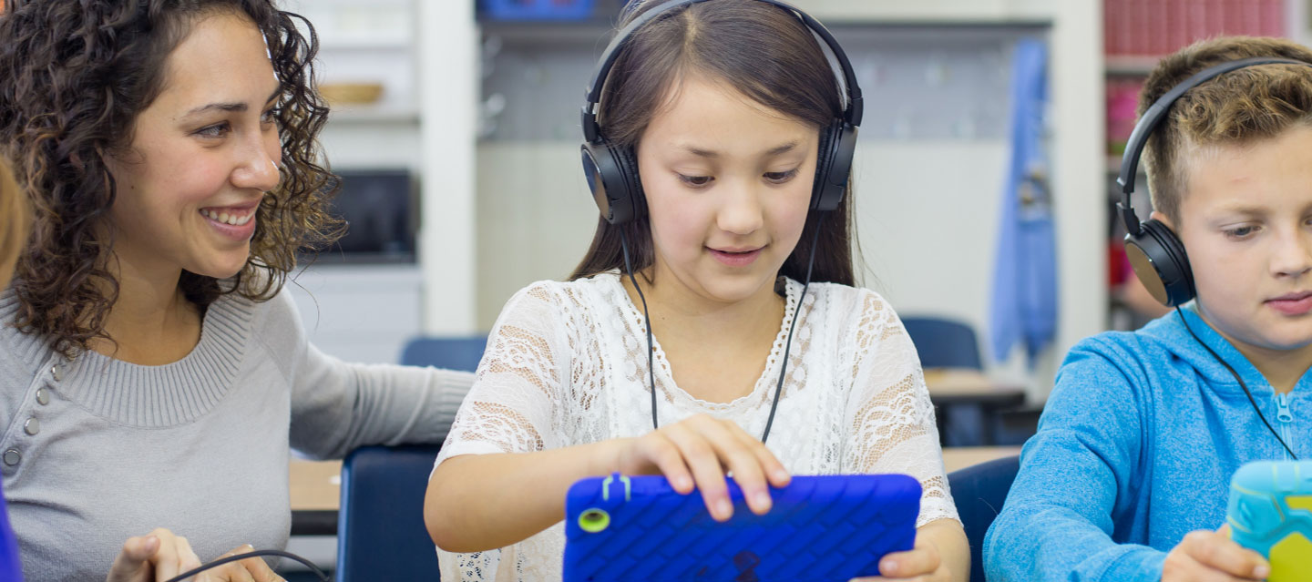 two elementary ed students with headphones on their laptops with teacher looking on