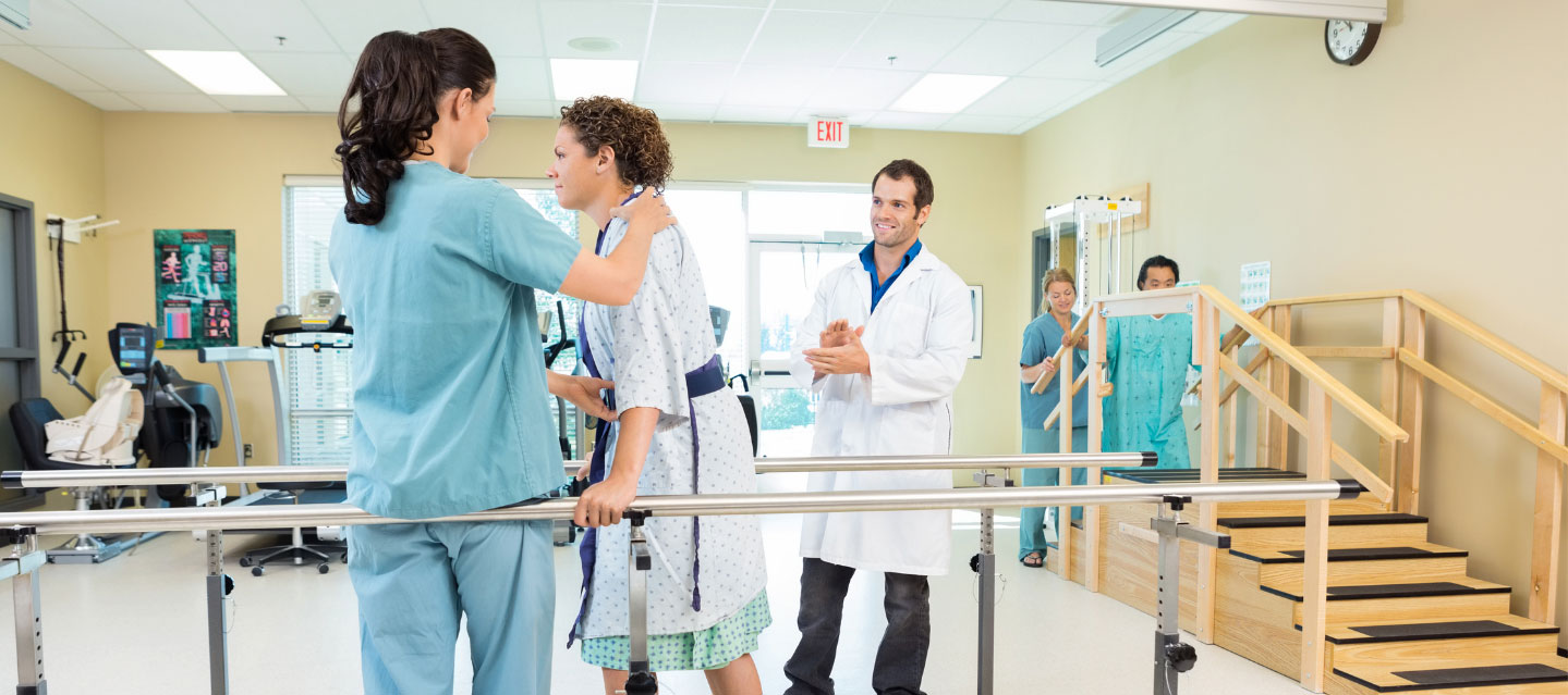 physical therapist helping patient to walk while holding on to bars.