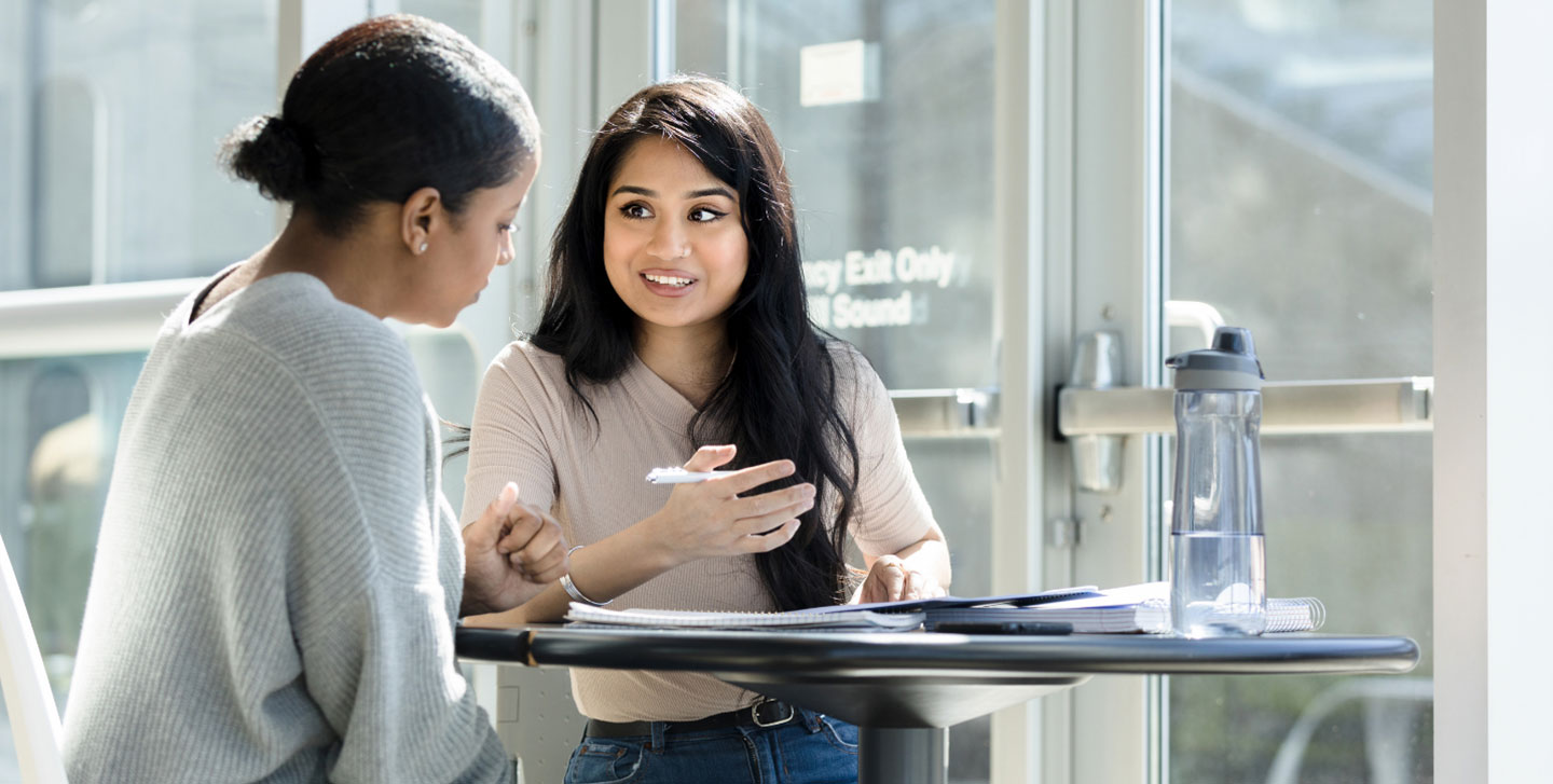 two female students sitting at table talking in atrium of Goodwin hall