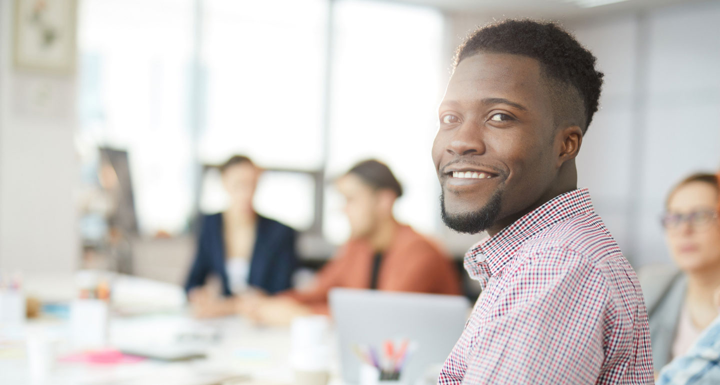 male student at table turning to face camera smiling, people in background blurred out; Entrepreneurship program
