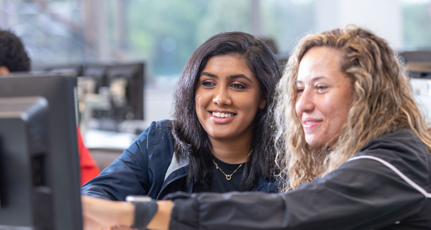 two female student working at a computer, one pointing to the monitor; marketing