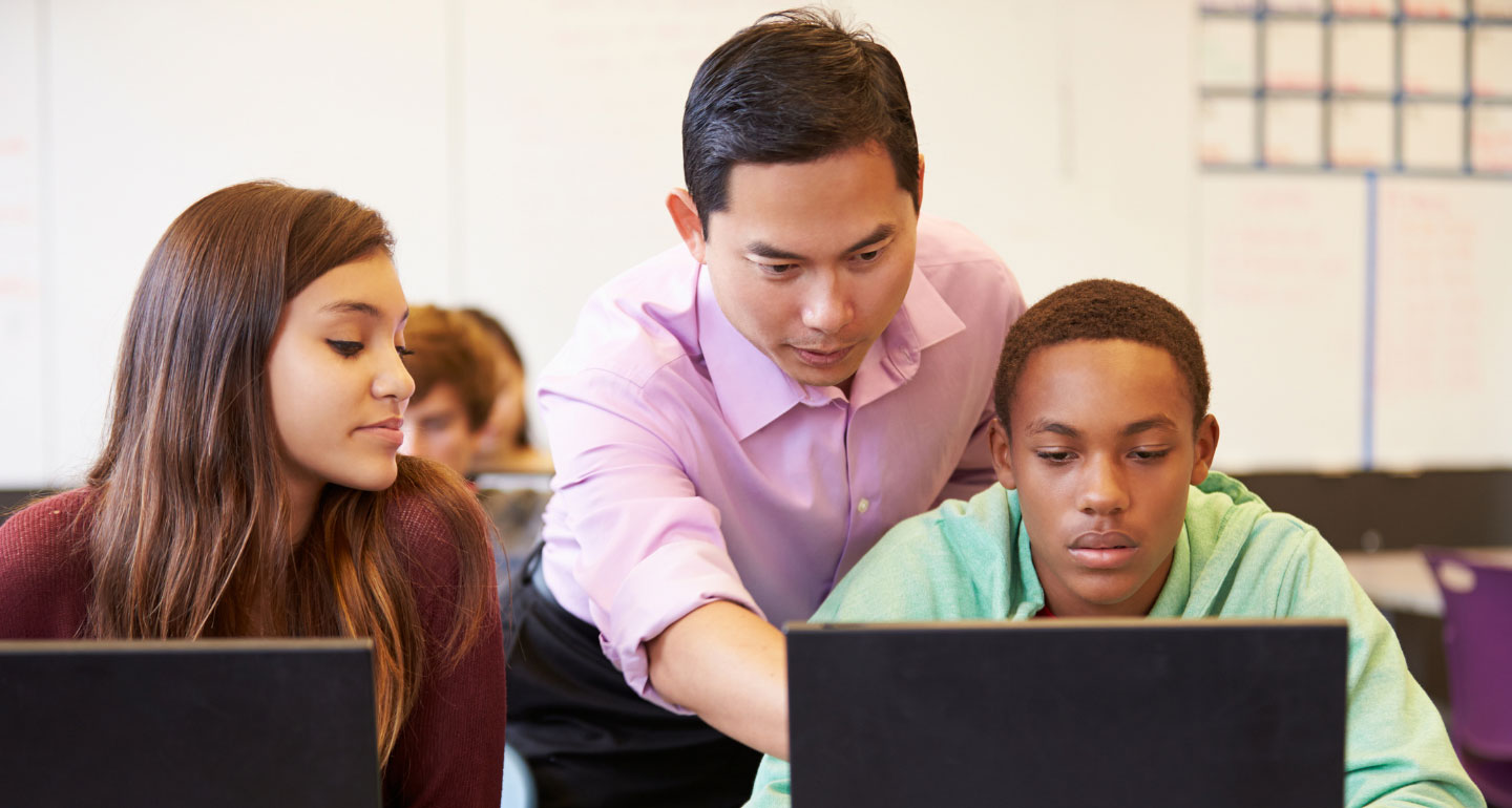 two students at computer station, male teacher point to one of the screens; secondary education program
