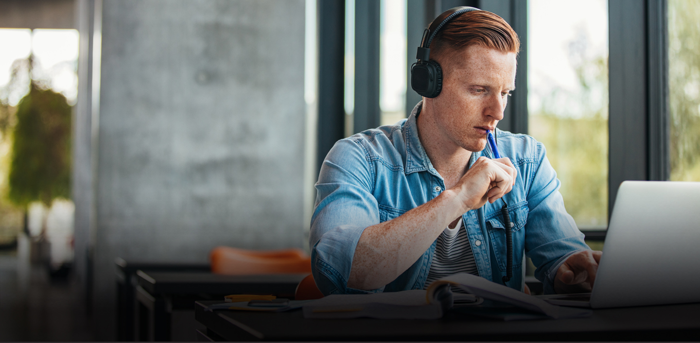 Male student wearing headphones holding a pen looking at his computer; Library Technology