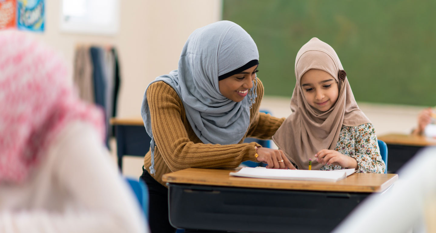 Indian female teacher instructing a young Indian female student, both with head scarfs, smiling