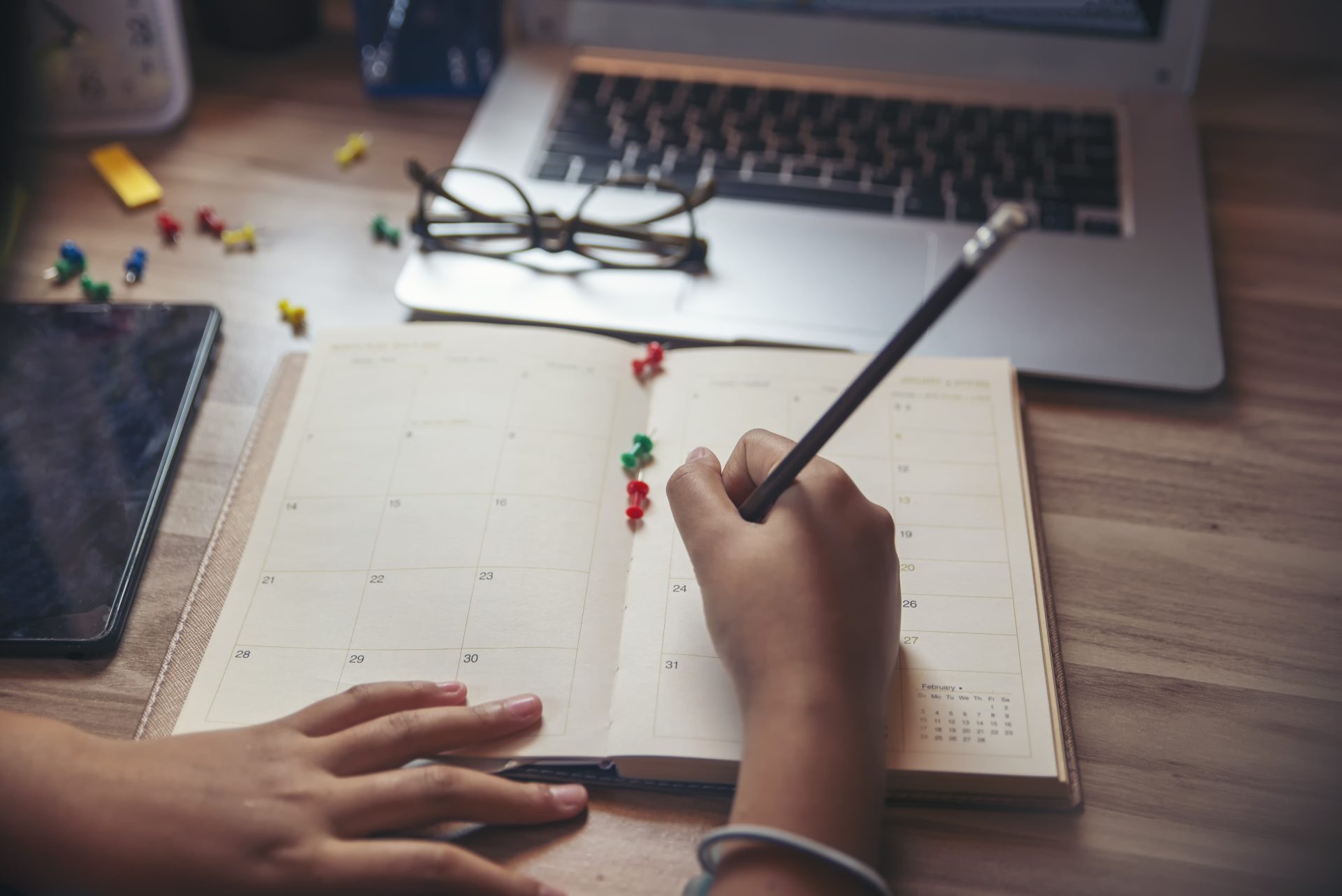 Hands writing in a calendar with a pencil, thumbtacks are scattered on the calendar and table - working from home