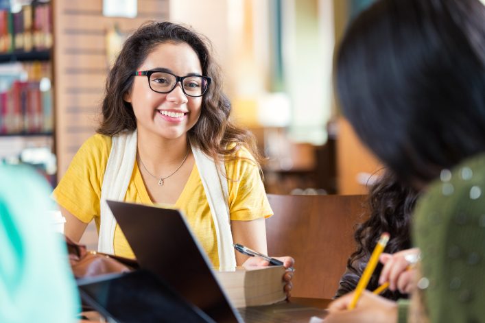 Trendy hipster Hispanic college age girl is smiling while sitting at tabe in library with group of friends. Students are working in study group to prepare for exam while using laptop computers, digital tablets, and other technology.