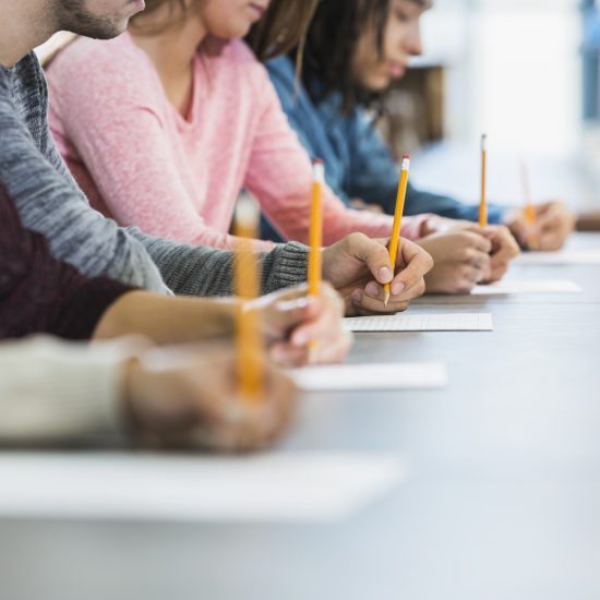 Cropped view of a multiracial group of young men and women sitting in a row at a table, writing with pencils on paper. They are taking a test or filling out an application. Focus is on the hand of the young man in the middle in the gray shirt.