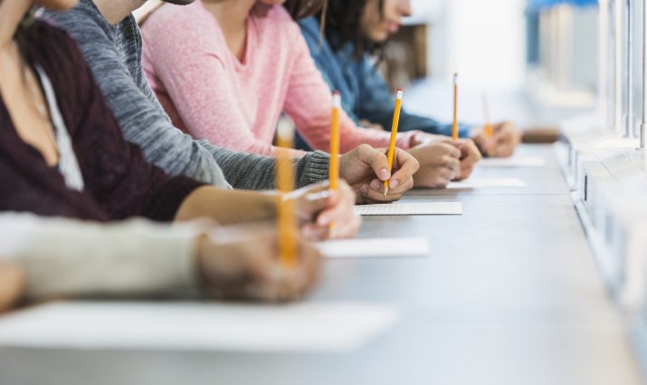 Cropped view of a multiracial group of young men and women sitting in a row at a table, writing with pencils on paper. They are taking a test or filling out an application. Focus is on the hand of the young man in the middle in the gray shirt.