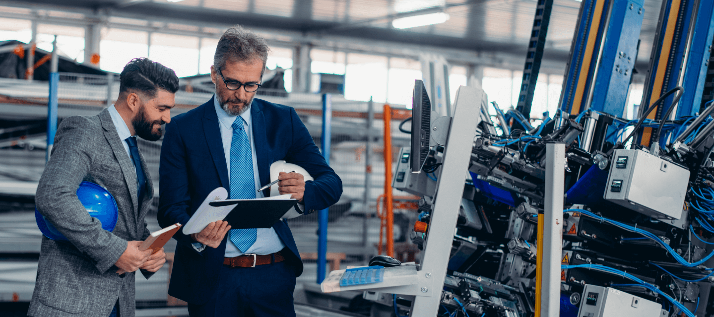 boss looking over clipboard of information with employee with machinery in the area - management